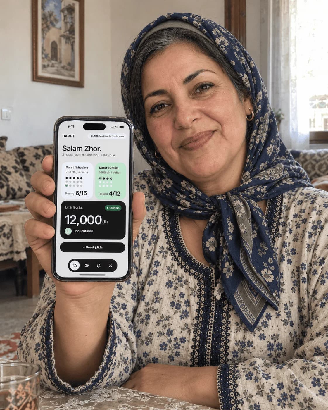 A woman in a patterned headscarf at a table, holding up a phone showing the Daret home screen.